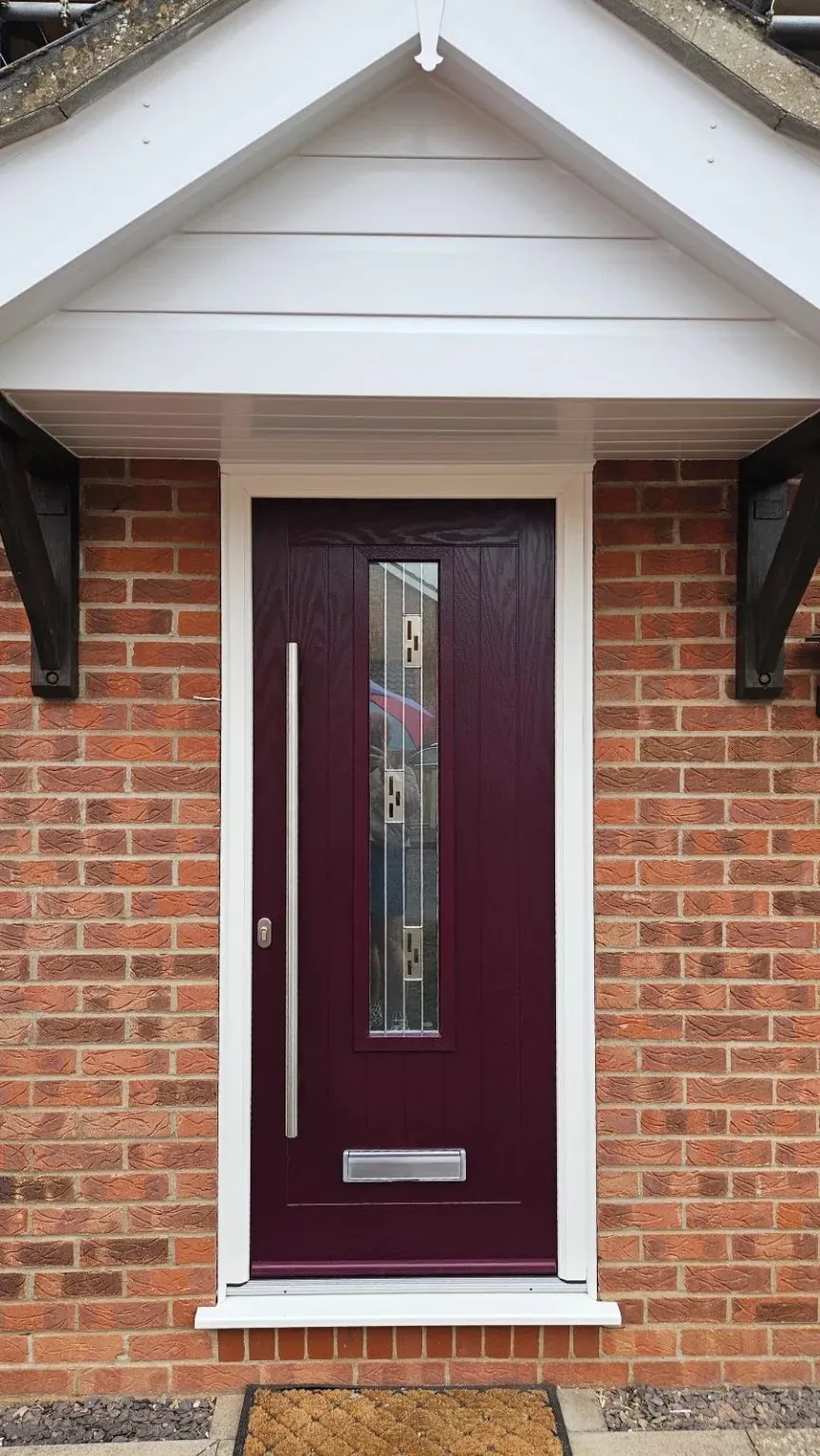 Deep red composite front door with a tall decorative glazed panel and long bar handle, in a white frame under a white clad porch, Drayton NR8