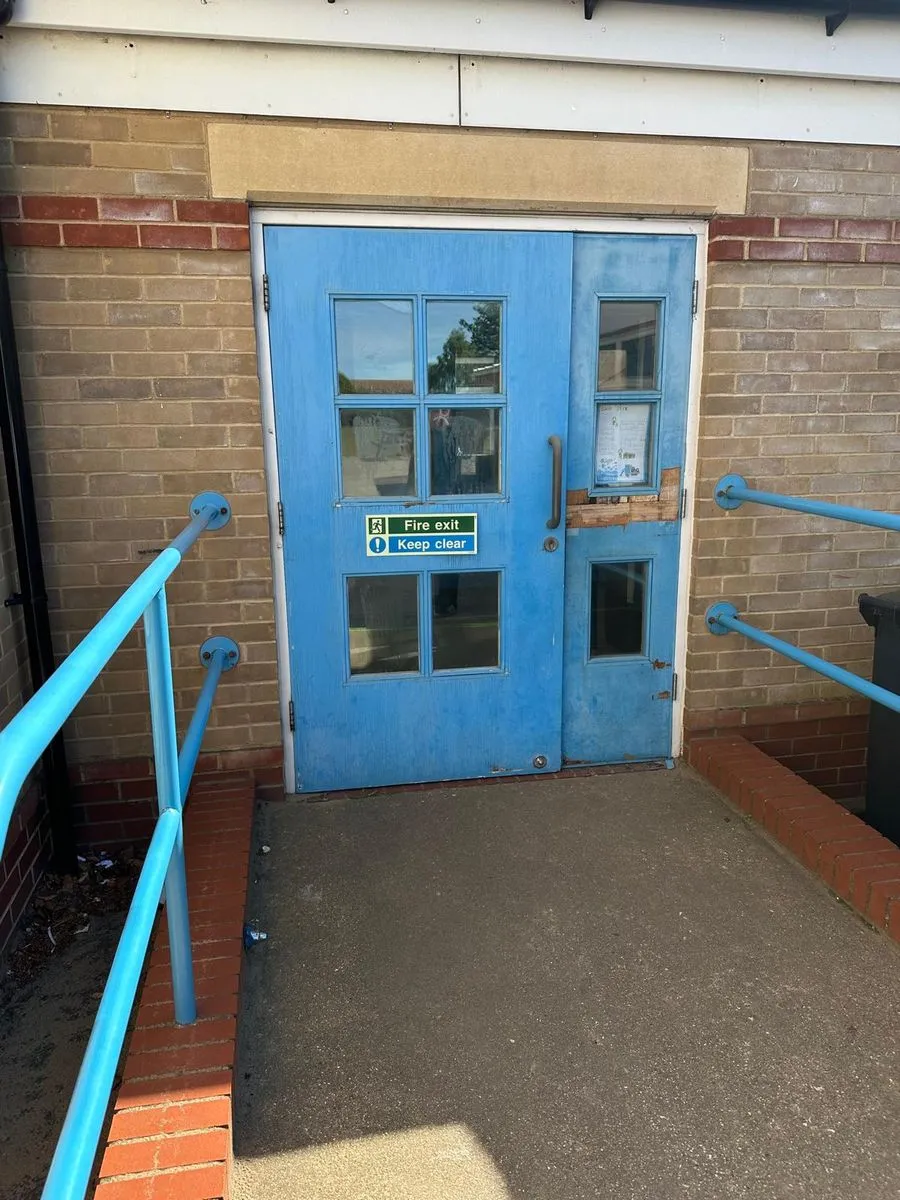Faded blue timber fire exit door with a boarded-up side panel at the top of a ramp at Horsford CofE VA Primary School